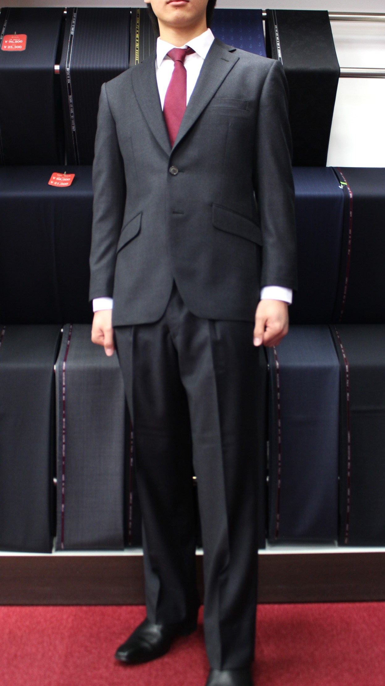 店内でスーツ生地が並ぶ棚の前に、成人男性がカノニコのグレーのスーツとエンジ色のネクタイを着用して正面を向いて立っている。表情は落ち着いており、全身が写っている。