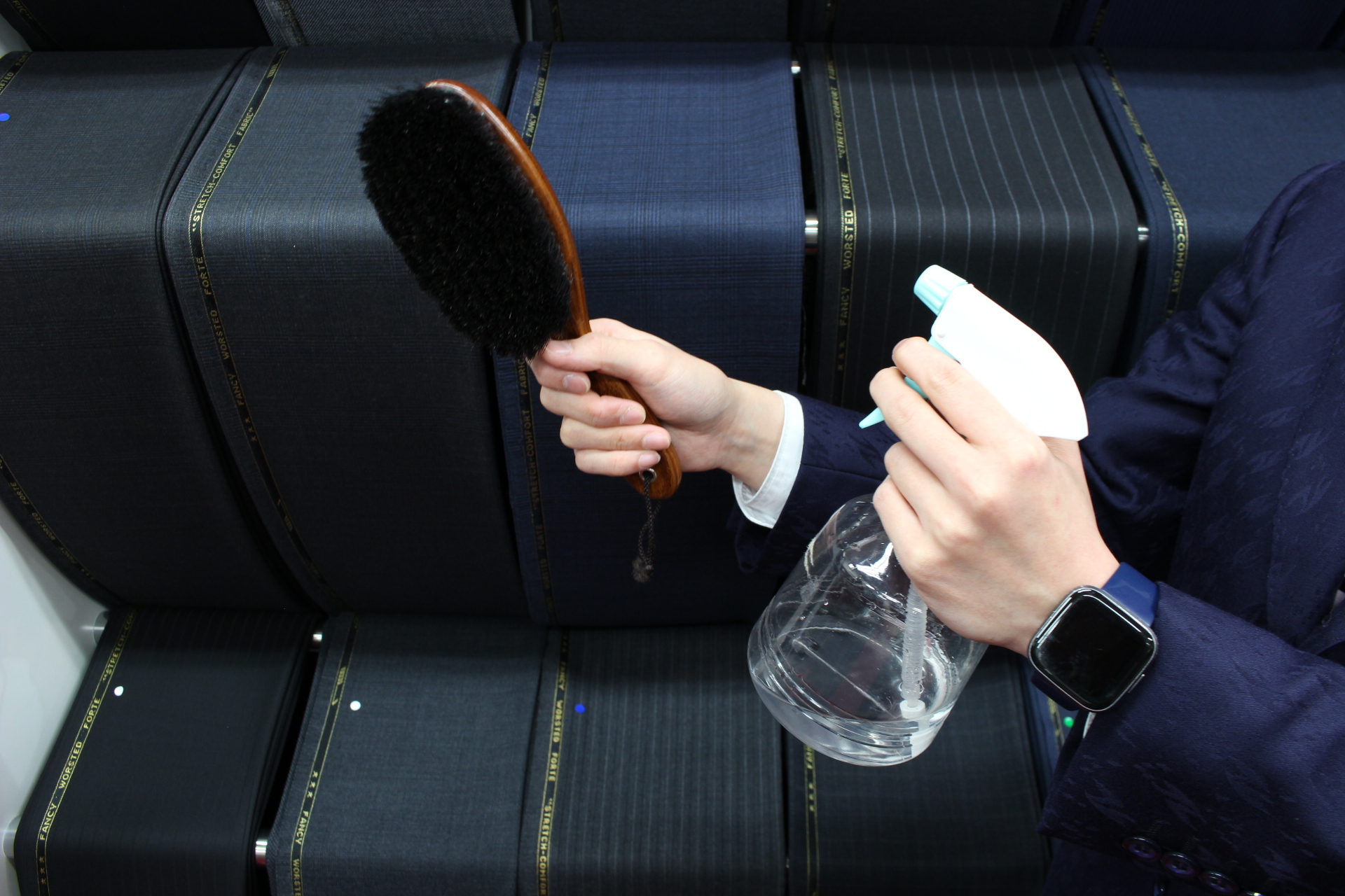 スーツ生地を背景に、洋服ブラシと霧吹きを両手に構える手元のアップ。衣類ケアの基本道具の紹介。