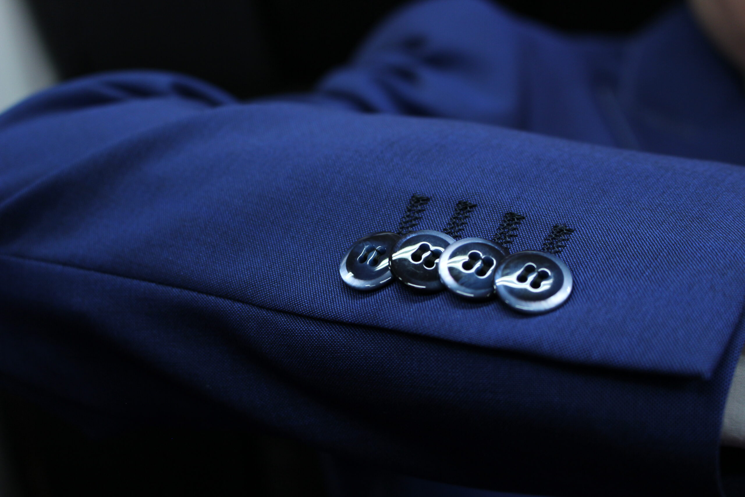 鮮やかなブルーのスーツの袖口（カフ）のアップ写真。光沢のあるダークネイビーのボタンが4つ、わずかに重なり合うように並べて縫い付けられた「重ねボタン」仕様のデザイン。