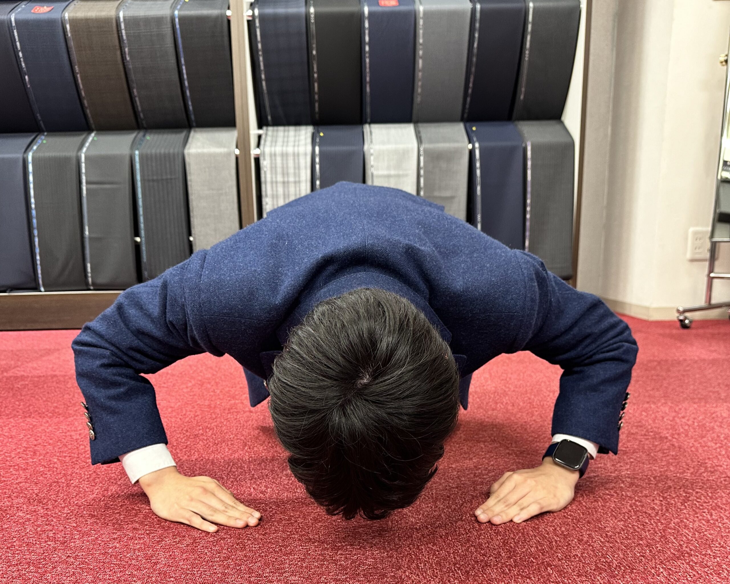 赤いカーペットの上で、青いジャケットを着た人物が深く土下座をしている俯瞰写真。背景にはさまざまな色や柄のスーツ生地のロールが棚に並んでいます。人物は左手首にスマートウォッチを着用しており、手はハの字の形で床についています。