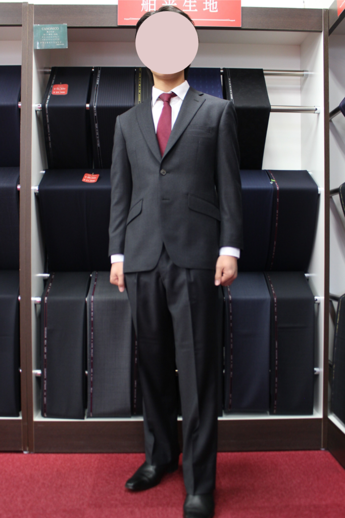 グレーの無地スーツを着用した男性の全身写真。イタリアの生地ブランド「カノニコ(CANONICO)」の生地を使用したシングル2つボタンのスーツで、インナーには白シャツとエンジ色のネクタイを着用。背景には紳士服店のスーツ生地が陳列された棚があり、落ち着いたビジネススタイルのコーディネートを紹介している。