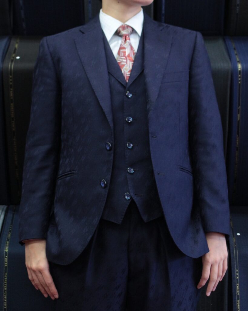 光沢感のあるネイビーの地模様(シャドージャガード)が施されたスリーピーススーツを着用した男性のバストアップ写真。シングルジャケット、5つボタンのベスト、スラックスの組み合わせ。白のワイシャツに、赤と白の幾何学模様がデザインされたネクタイを合わせている。ボタンは光沢のあるシェルボタン風のものを採用しており、華やかでドレッシーな印象のコーディネート。