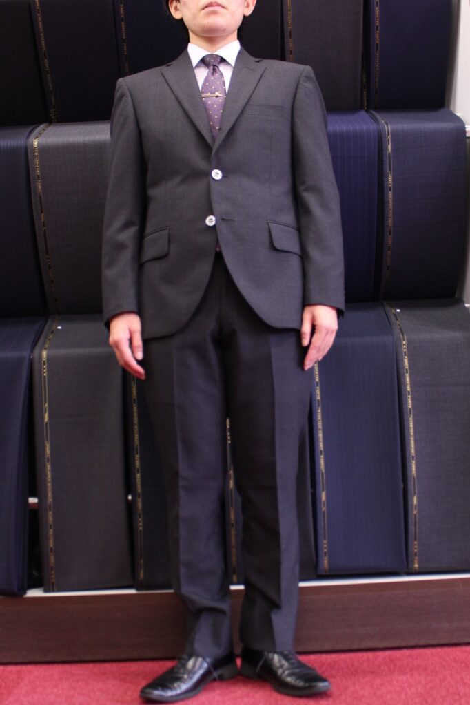 ダークグレーのスーツを着用し、直立した男性の正面全身写真。スーツのジャケットは2つボタンを留めており、紫色のネクタイにネクタイピンを着用しています。背景にはスーツ生地(反物)のロールが整然と並んでおり、仕立て屋やスーツ専門店の店内のような風景です。